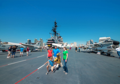 The deck of the U.S.S. Midway Aircraft Carrier which has several military aircraft parked on top of it, several visitors to the ship walking around sightseeing and the buildings of downtown San Diego peering out in the distance